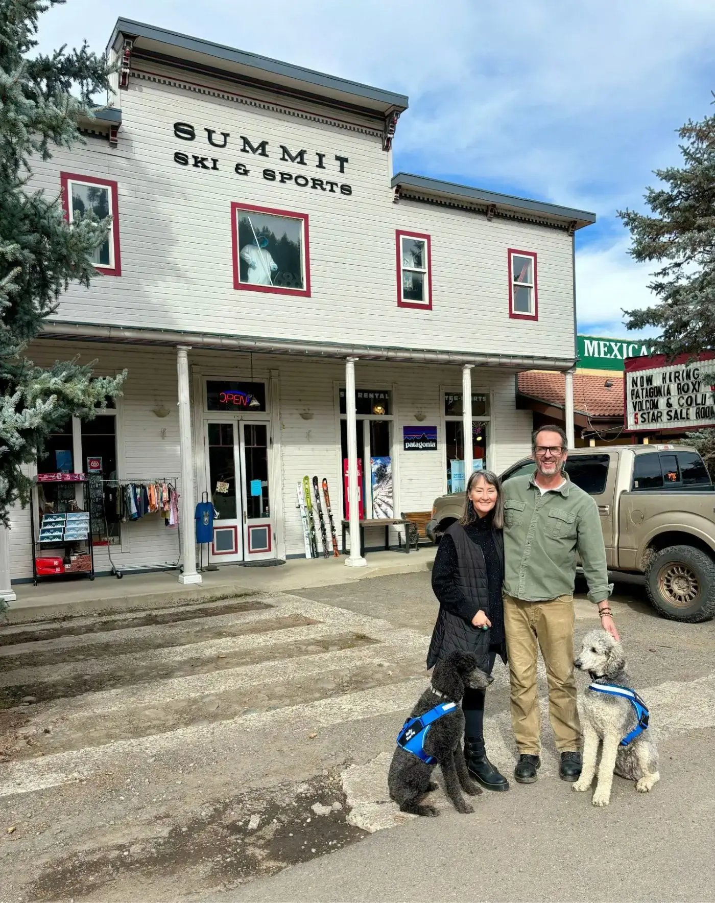 Two people with their dogs stand outside the Summit Store & Post in a small town.
